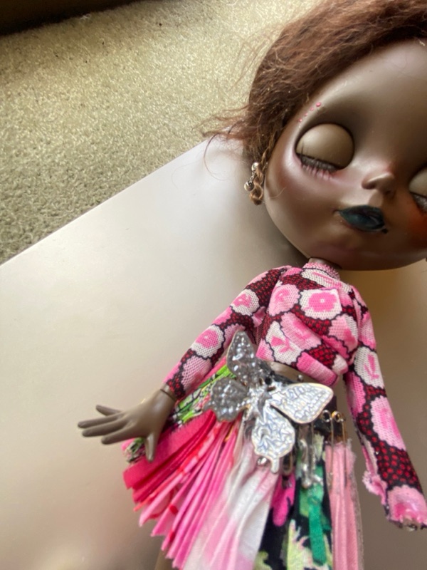 Cait's custom Blythe: matte choco face mould, auburn hair with small braids, eyes closed, teal lips, in a pink print top and a pleated pink skirt with a silver butterfly